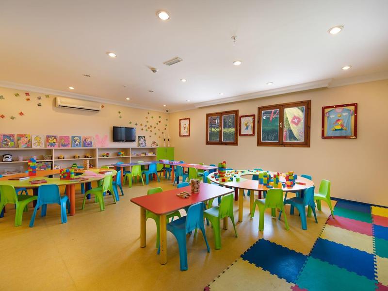 Sala giochi colorata con piccoli tavoli e sedie, decorazioni murali e tappeto da gioco.