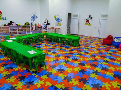 Sala giochi colorata con tavoli verdi, pavimento con tappeti puzzle colorati e pouf in un angolo.