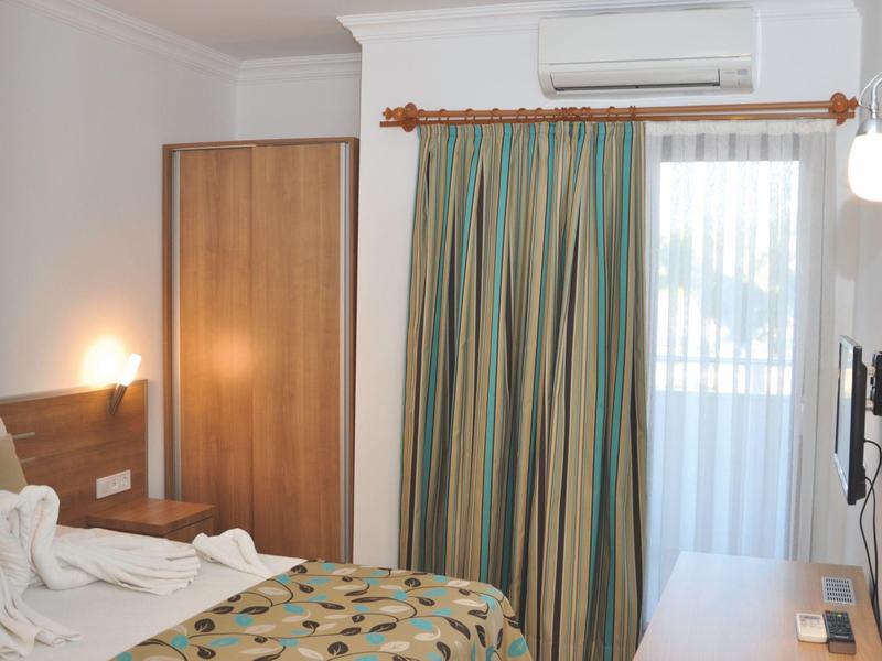 Helles Hotelzimmer mit Bett, Holzschrank, bunten Vorhängen und Balkonzugang.