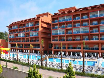 Großes rotes Hotelgebäude mit mehreren Balkonen, großem Pool und Sonnenschirmen im Vordergrund.