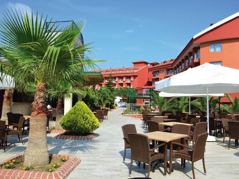 Area esterna di un hotel con sedute, ombrellone e palme sotto un cielo azzurro.