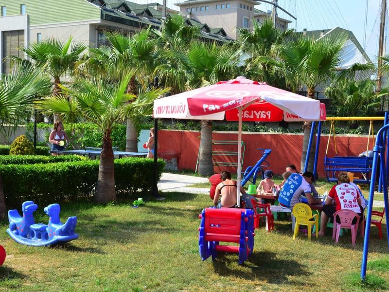 Parco giochi con attrezzature colorate, ombrelloni rossi e palme sullo sfondo.