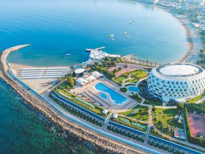 Luftaufnahme einer Küstenanlage mit weißem, rundem Gebäude, Pool und Promenade am Meer.