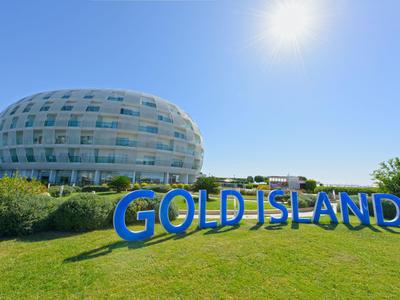 Rundes modernes Gebäude hinter einem grasbewachsenen Hügel mit blauer "GOLD ISLAND"-Beschriftung.