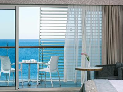 Balkon mit Glasgeländer zeigt zwei weiße Stühle, Tisch und Meer im Hintergrund, Blick aus Zimmer.