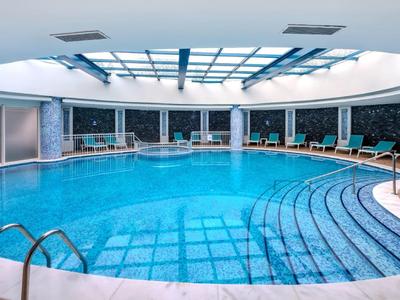 Piscine intérieure ronde éclairée en bleu avec chaises longues et toit en verre dans un espace bien-être moderne.