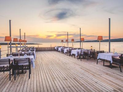 Buitentafels met witte tafelkleden en stoelen op een houten terras aan het water bij zonsondergang.