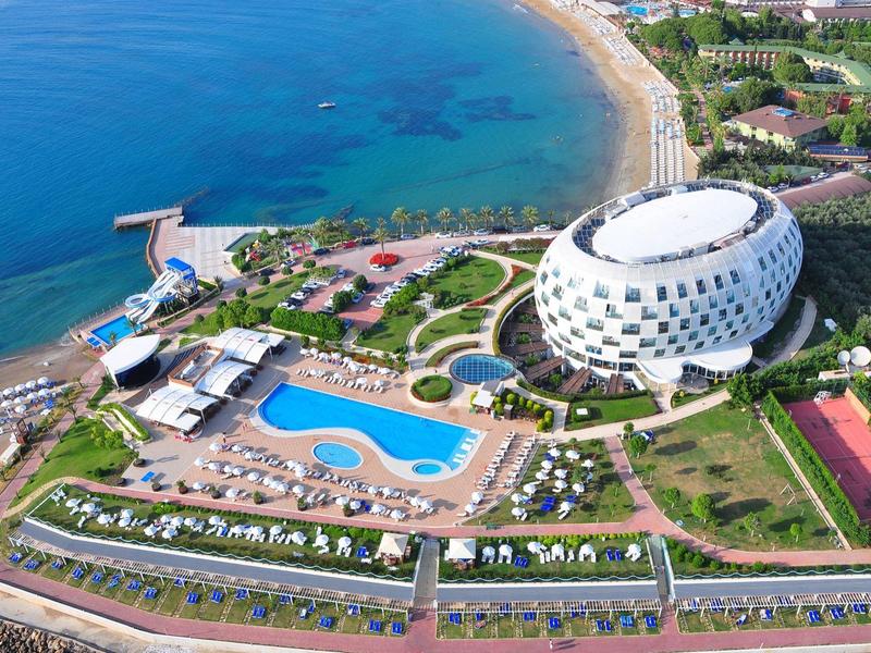 Vue aérienne d'un hôtel moderne en forme ovale avec piscine, plage et mer bleu clair.