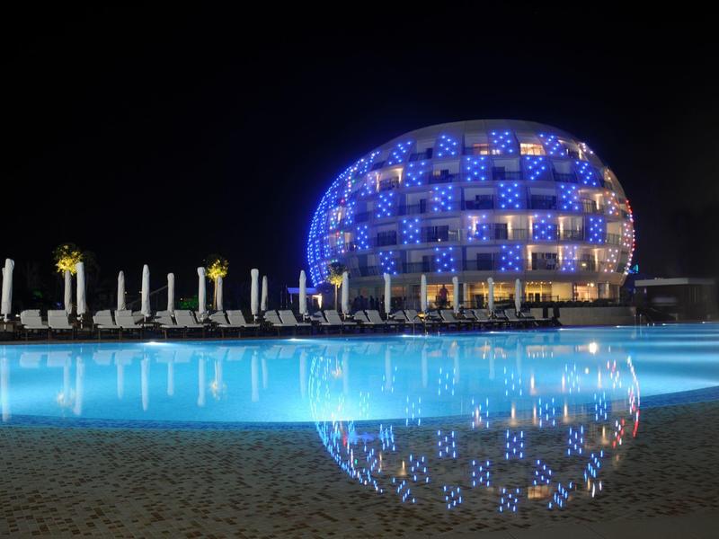 Bâtiment moderne rond illuminé bleu la nuit, reflet dans piscine extérieure calme.
