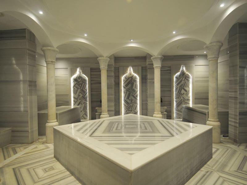 Salle de bain hammam avec marbre beige, lavabos encastrés et miroirs éclairés.