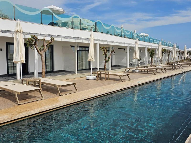Piscine longue devant une rangée de transats et parasols blancs sous un ciel bleu.