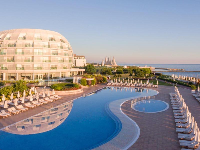 Grande piscine devant un hôtel moderne avec vue sur la mer au coucher du soleil.