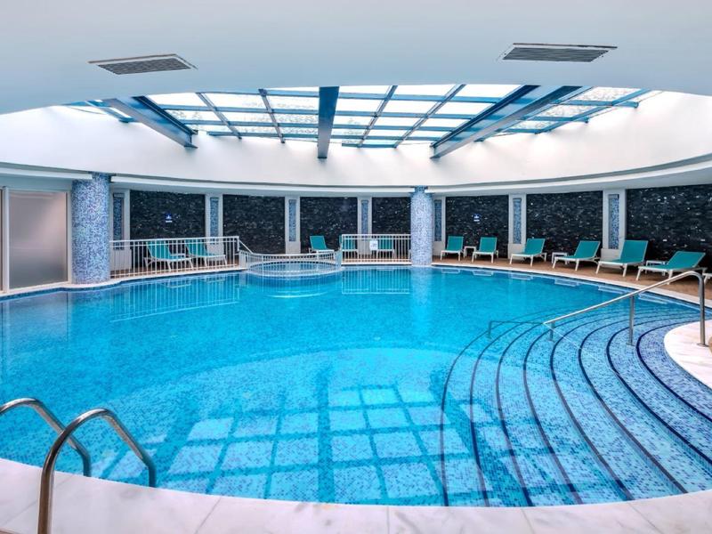 Piscine intérieure ronde éclairée en bleu avec chaises longues et toit en verre dans un espace bien-être moderne.