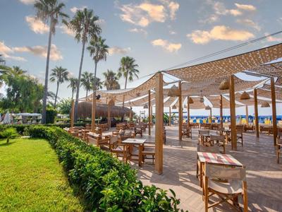 Offenes Restaurant mit Holztischen und Stühlen, Palmen, grünem Rasen und blauem Himmel am Meer.