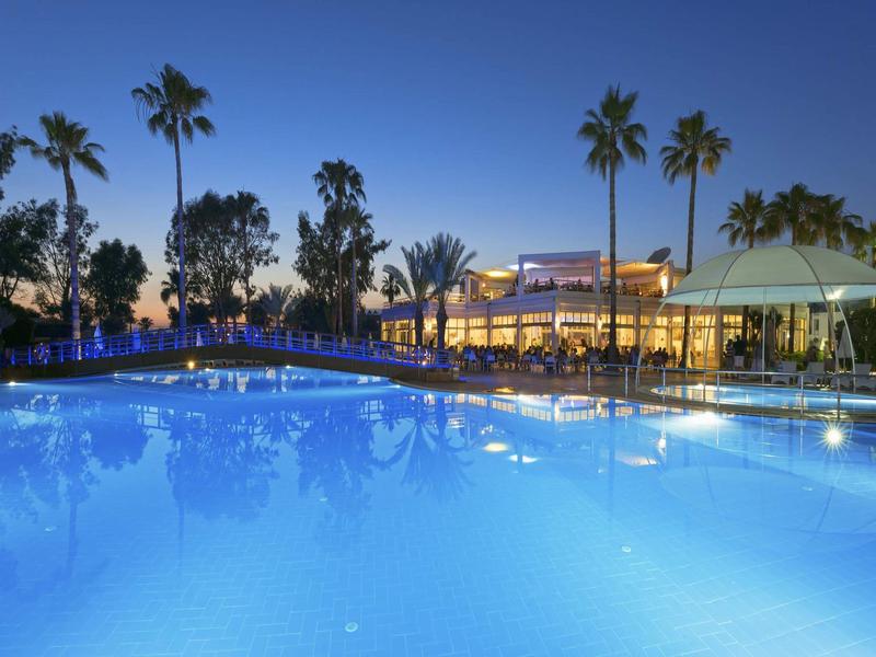 Piscina de hotel iluminada al anochecer con palmeras y casa de huéspedes al fondo.