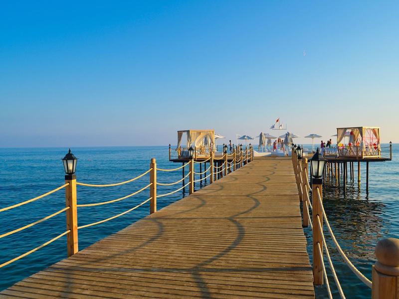 Largo muelle de madera se extiende sobre el mar hacia pequeños bungalows sobre el agua bajo un cielo despejado.