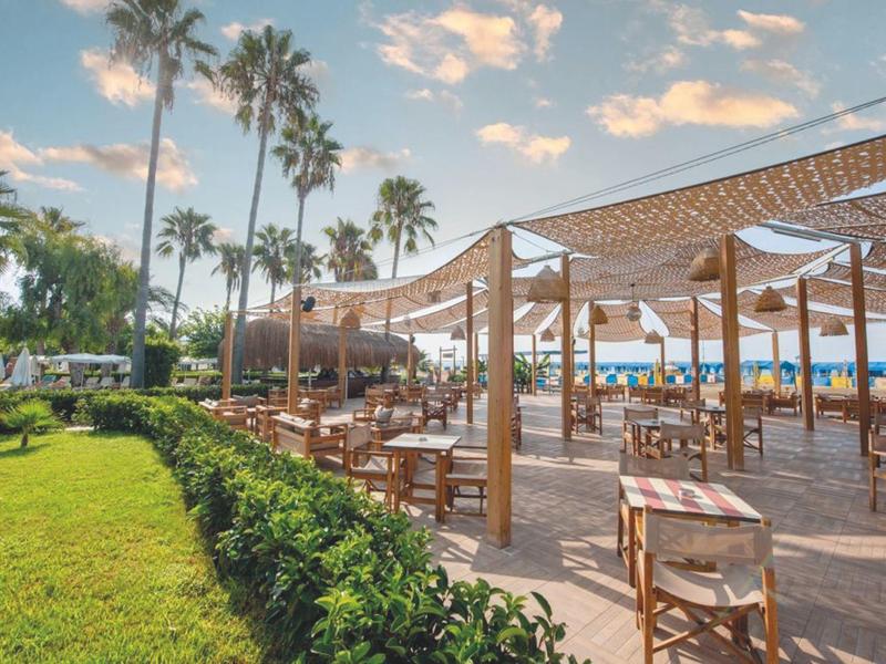 Zona de restaurante al aire libre con sillas y mesas de madera bajo velas, palmeras y mar al fondo.