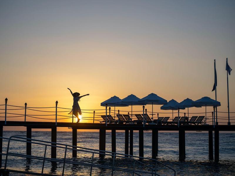Una persona salta alegremente en un muelle al atardecer junto al mar con sombrillas de fondo.