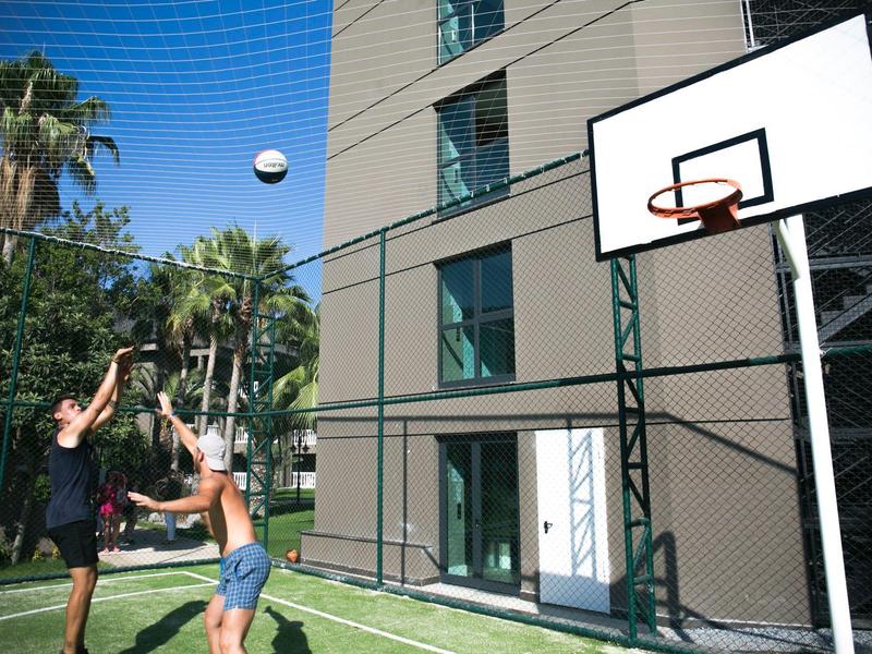 Dos personas juegan baloncesto en una cancha al aire libre junto a un edificio de hotel bajo un cielo azul.
