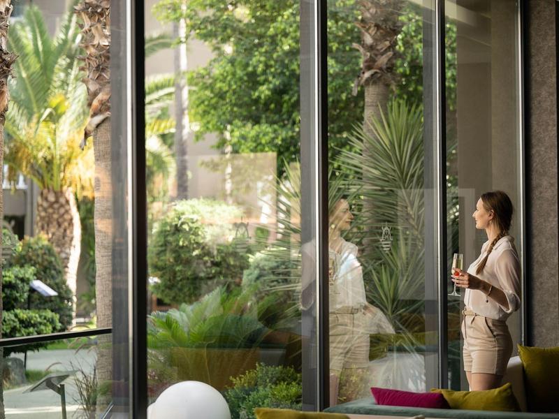 Mujer disfruta café mirando un jardín verde a través de grandes ventanales en un hotel.