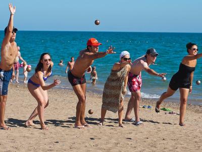 Sieben Personen spielen Strandvolleyball am Meer, Sand unter den Füßen, blaue Wellen im Hintergrund.