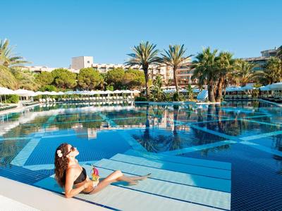 Frau entspannt am großen Hotelpool mit Palmen und weißen Sonnenschirmen bei Sonnenschein.