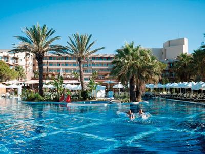 Blauer Pool mit Palmen, Sonnenschirmen und Hotelgebäude bei sonnigem Himmel.