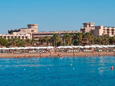 Großer Hotelkomplex mit mehreren Stockwerken, Palmen und vielen weißen Sonnenschirmen am blauen Meer.