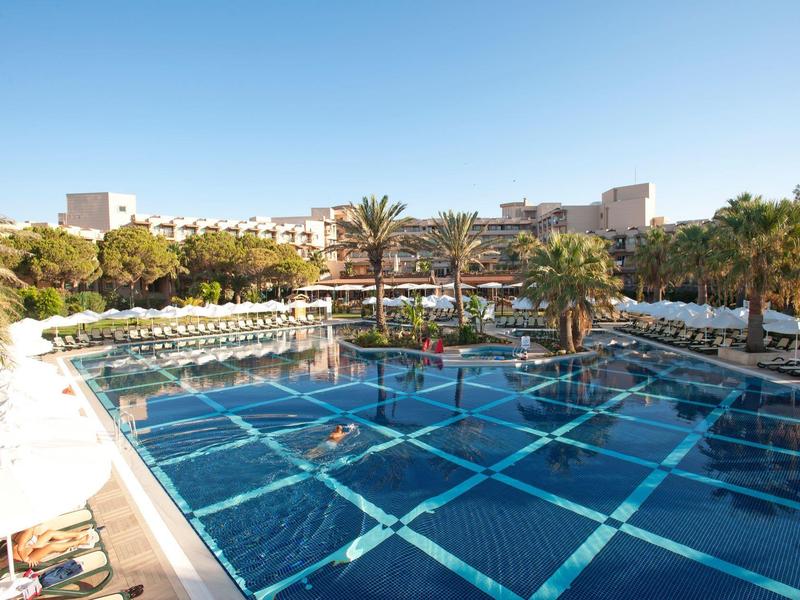 Large hotel pool with sun umbrellas and loungers, palm trees, and hotel buildings in the background.