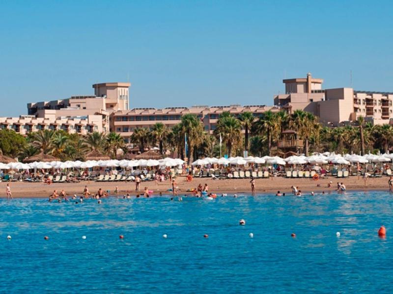 Large beachfront hotel with palm trees and umbrellas by turquoise sea.