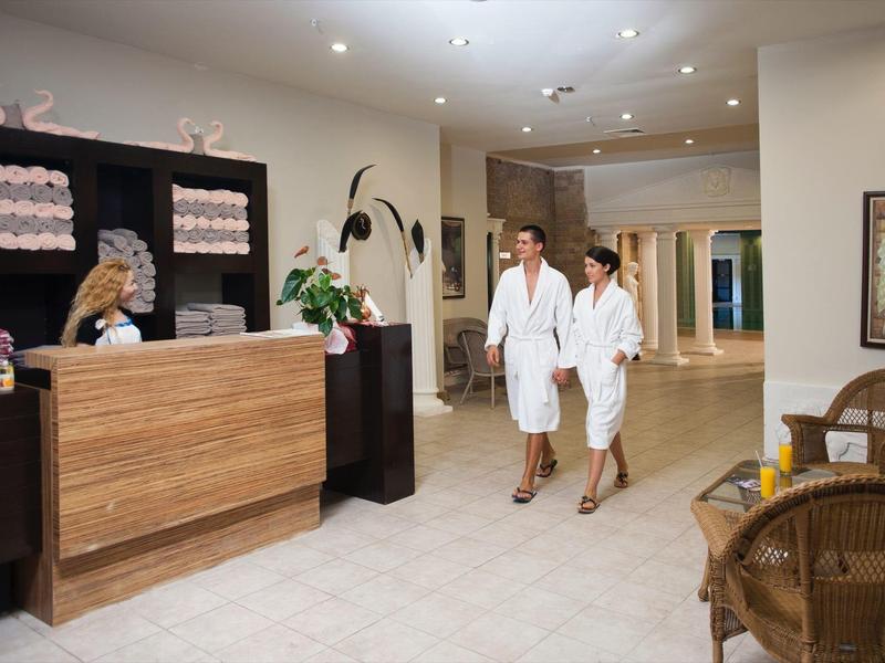 Hotel reception with a receptionist, stacked towels, and guests in bathrobes.