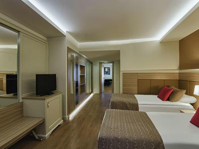 Moderní hotelový pokoj se dvěma postelemi, TV, skříní a nepřímým osvětlením na stropě.