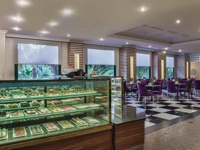 Elegantní kavárna se skleněnými vitrínami plnými pečiva a fialovými židlemi ve velkém prostoru.