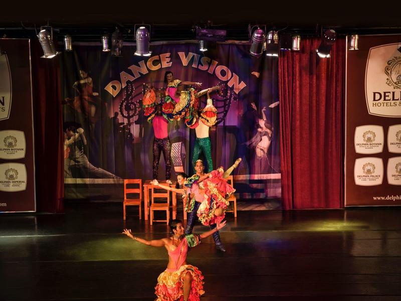 Dva tanečníci v barevných kostýmech předvádějí živé vystoupení na jevišti s červenými závěsy a transparentem 'Dance Vision'.