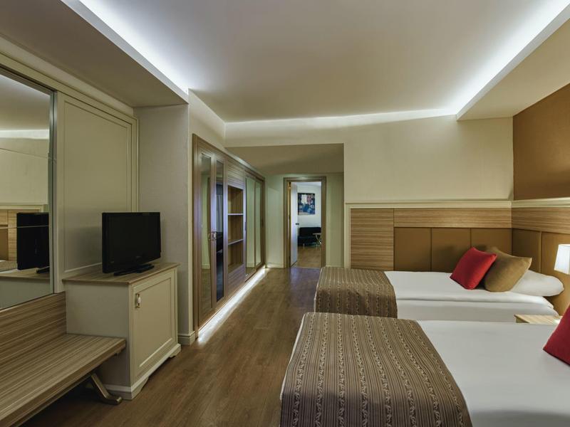Moderní hotelový pokoj se dvěma postelemi, TV, skříní a nepřímým osvětlením na stropě.