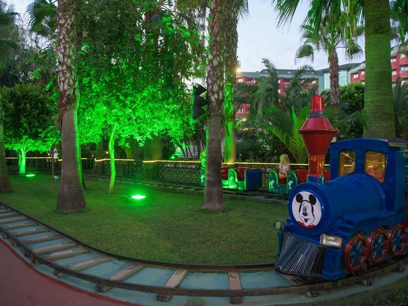 Malý dětský vlak s modrou lokomotivou na hřišti pod palmami a zeleným osvětlením.