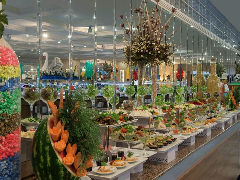 Dlouhý bufetový stůl s různými pokrmy a květinovými aranžmá v slavnostně vyzdobeném prostoru.