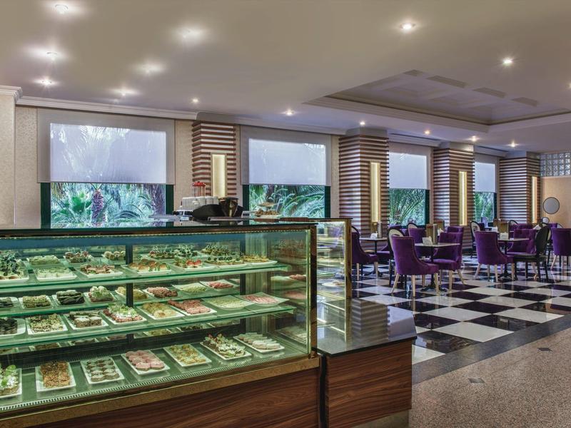 Elegantní kavárna se skleněnými vitrínami plnými pečiva a fialovými židlemi ve velkém prostoru.