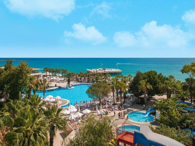 View of a large hotel pool with palm trees and sea view on a sunny day.