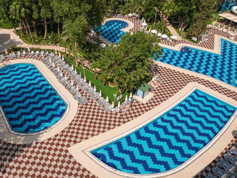 View of multiple zigzag-patterned pools surrounded by lounge chairs and trees in a hotel area.