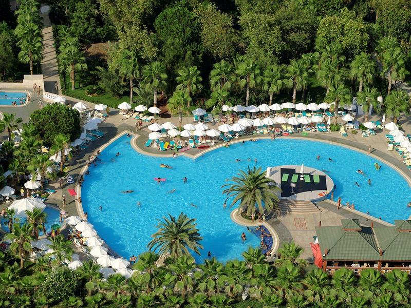 Large oval pool surrounded by sun loungers and lush greenery at a holiday resort.