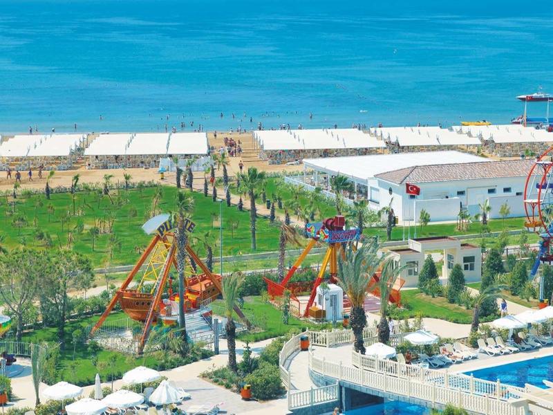 Veduta aerea di un parco divertimenti sul mare con ruote panoramiche colorate e giardini verdi.