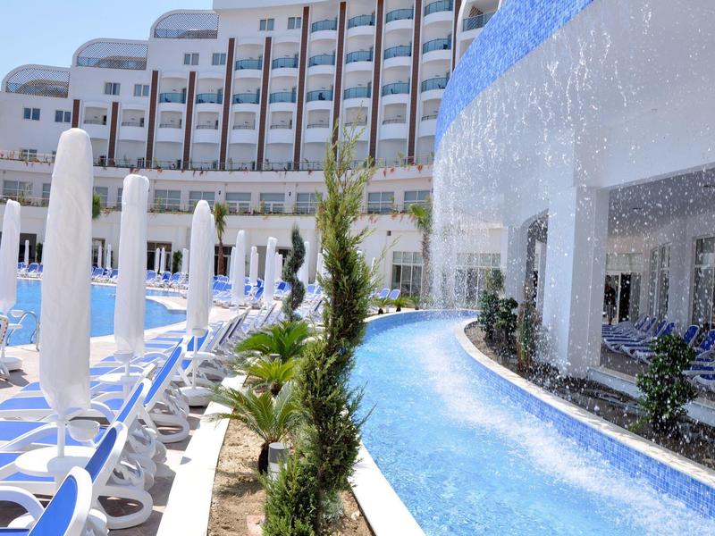 Area esterna di un hotel con piscina, lettini prendisole e giochi d'acqua sotto il sole.