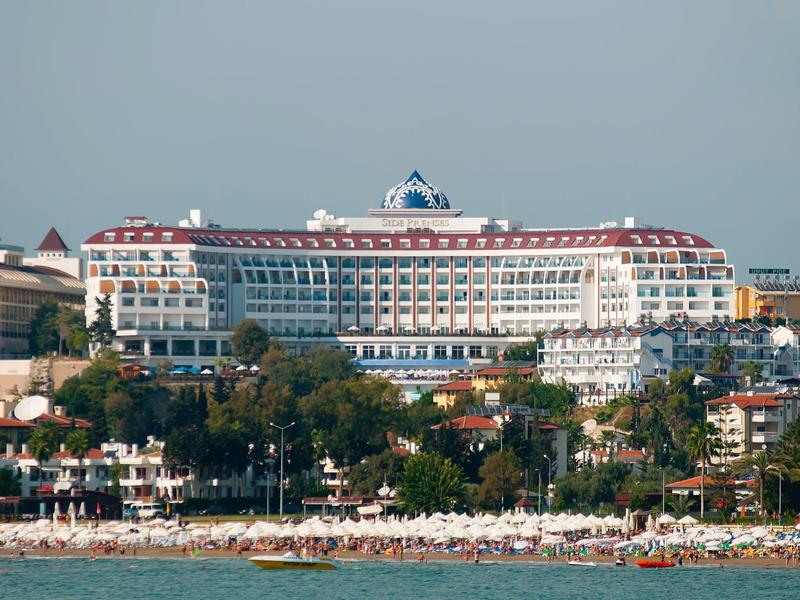 Grande edificio dell'hotel con tetto rosso dietro gli alberi e spiaggia vicino al mare.