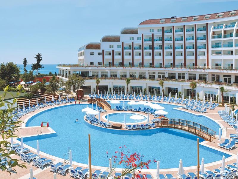 Grande piscina dell'hotel con lettini e ombrelloni davanti a un edificio su più piani.