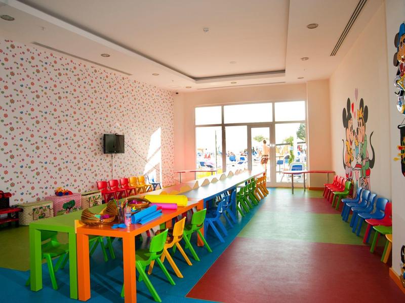 Sala giochi colorata per bambini con tavoli, sedie e decorazioni murali in una stanza luminosa con finestra.