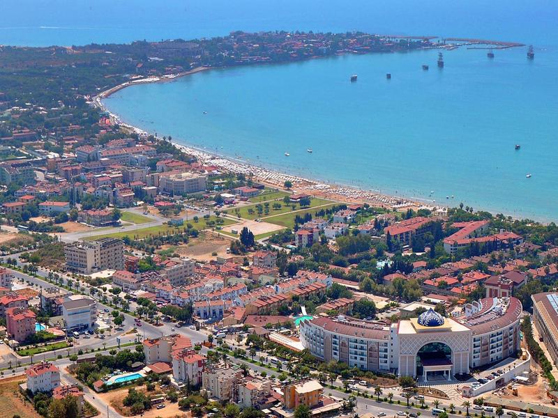 Veduta aerea di una città costiera con spiaggia, edifici e aree verdi con tempo sereno.