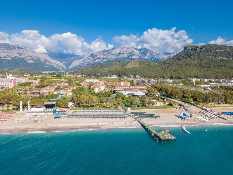 Vista di una spiaggia sabbiosa con un molo, terra verde sullo sfondo e montagne sotto un cielo blu.