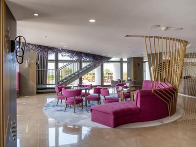 Area lounge moderna di hotel con sedie e divani rosa, pavimento chiaro e grandi finestre.