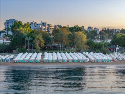 Plage avec transats blancs devant un parc vert et des bâtiments au coucher du soleil.